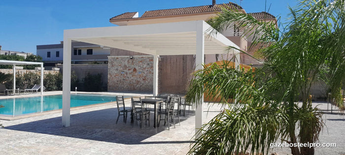 Pergola QBox Linea bianca accanto alla piscina, con tavolo e sedie sotto e una palma davanti