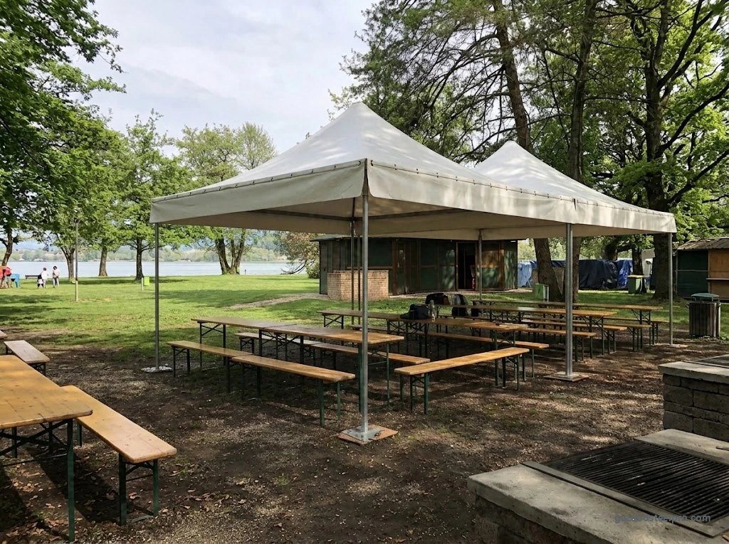 Due gazebo Corallo Light affiancati visti di lato, con tavoli e panche sotto, in un parco sul lago, foto vera